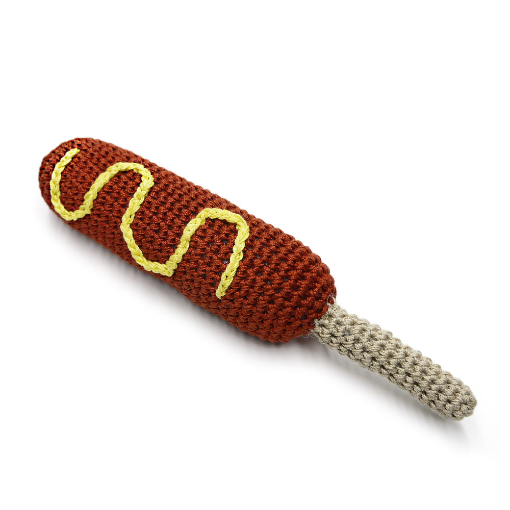 Crocheted hot dog toy with yellow wavy mustard stripes, dark brown body, gray string end
